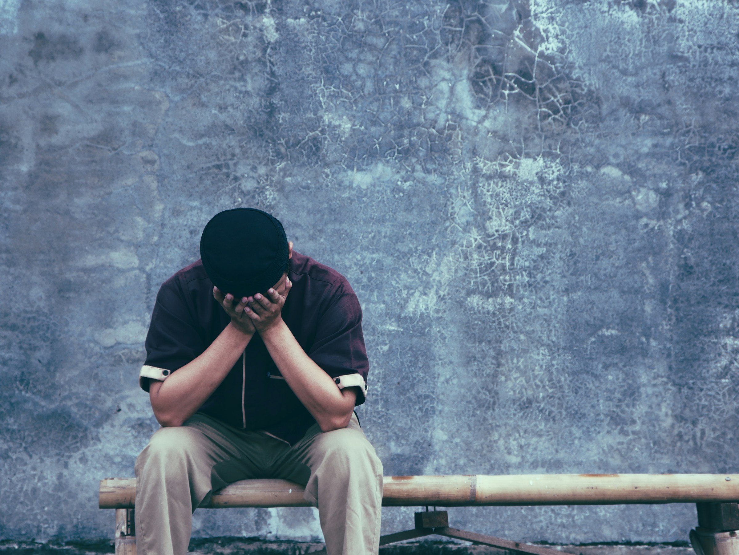 A man sitting on a bench outside with his head in his hands, thinking about burnout symptoms and depression he's experiencing.