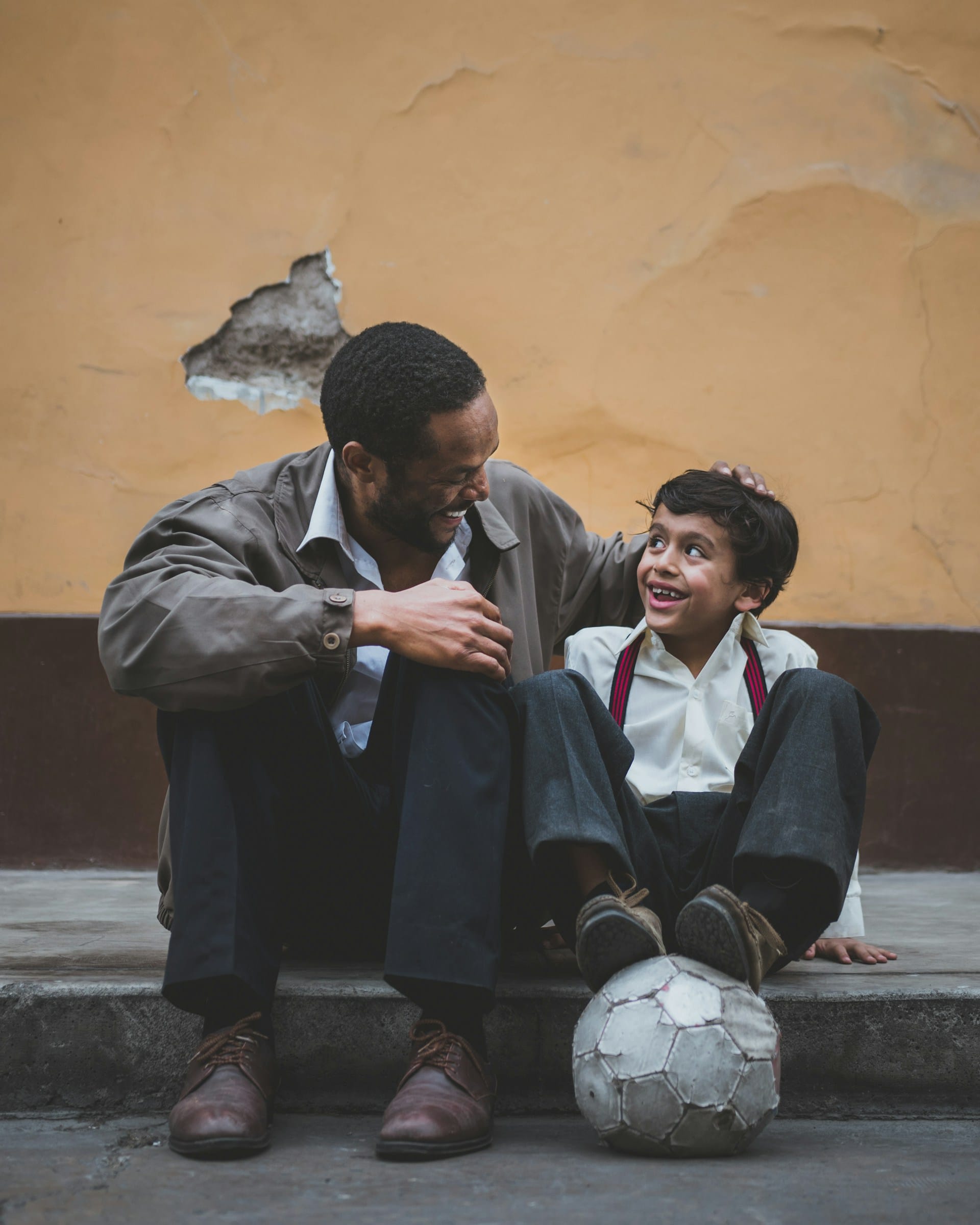 Father and son sitting on a curb with a soccer ball after the dad has talked with his friends about how to raise children in community together.