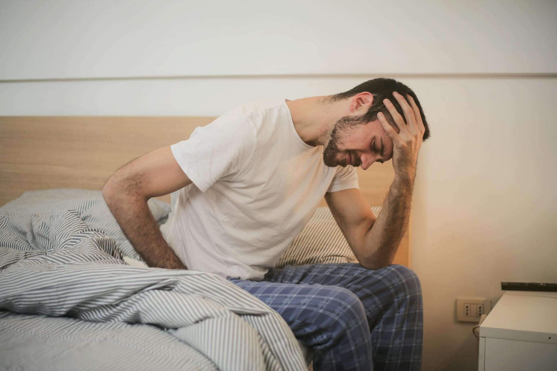 Man sitting on the side of his bed in pain, holding his back and his head, and trying to determine how to practice self-compassion.