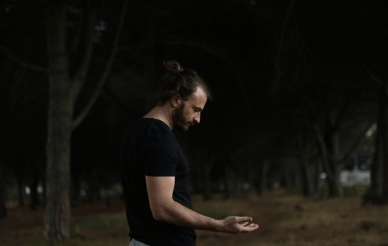 a man in a black t - shirt and jeans is sitting on the ground in front of a tree and looking at the camera